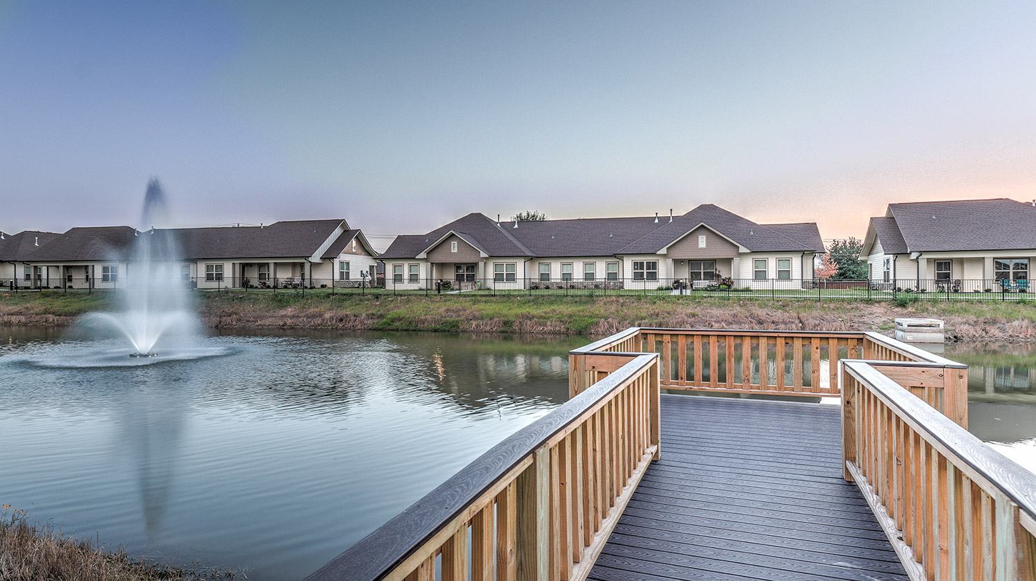 Peaceful fishing pier overlooking a pond at Ridgemere Senior Living, with water fountain, walking paths, and community homes in the background.