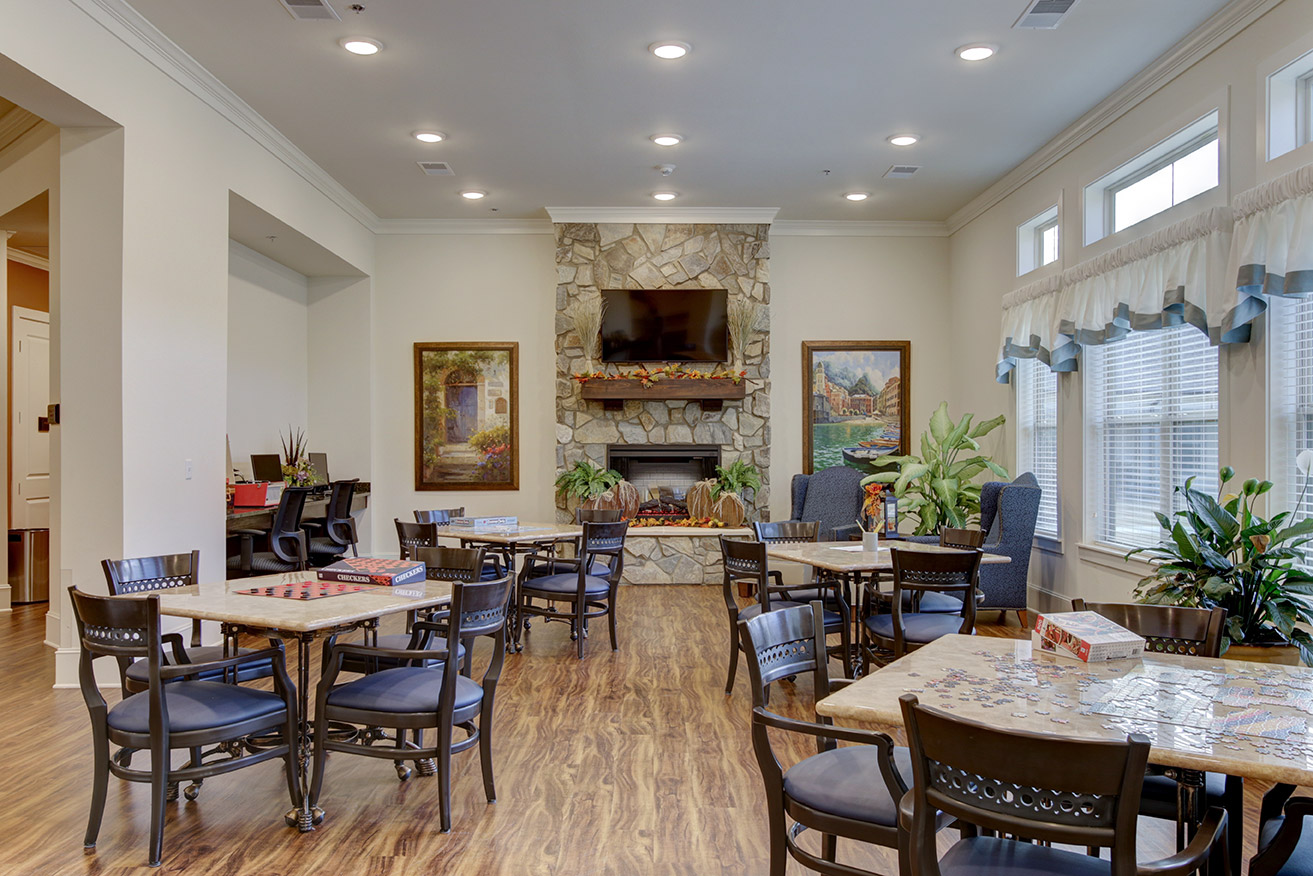 Game room at Ridgemere Senior Living featuring tables for puzzles and games, comfortable seating, natural light, and a stone fireplace creating a social gathering space.