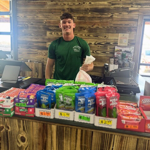 A young man smiles brightly behind the counter at a local business, surrounded by convenient snack options available for purchase.