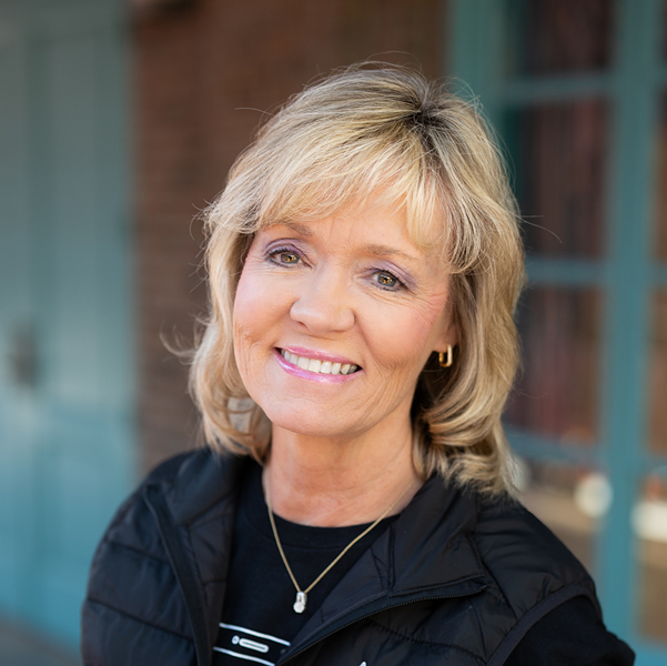 Sherri Newton, Executive Director at Ridgemere Senior Living, smiling in a professional outdoor headshot with short blonde hair, gold earrings, and softly blurred brick and teal window details in the background.
