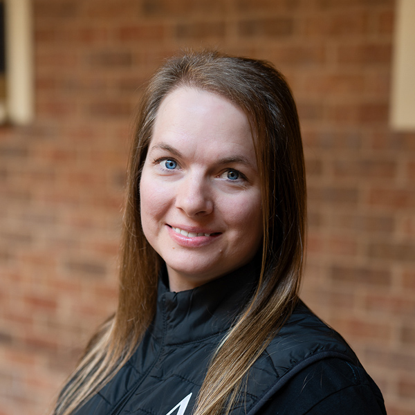 Ashley Thomas, Wellness Director at Ridgemere Senior Living, looking at the camera in a professional outdoor headshot with long straight brown hair, blue eyes, a black vest, and a softly blurred brick background.