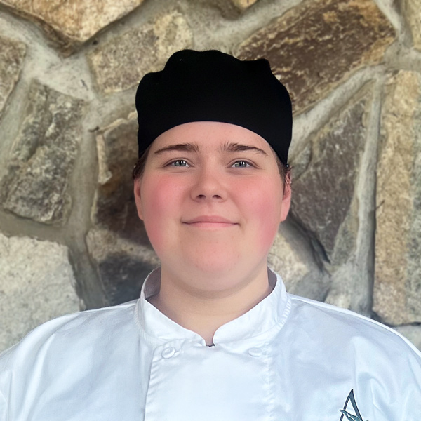 Camry Clark, Culinary Director at Ridgemere Senior Living, looking at the camera in a professional headshot wearing a white chef coat and black chef cap against a stone wall background.