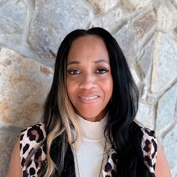 Emerald “Nisey” Newton, Memory Care Director at Ridgemere Senior Living, smiling in a professional headshot with long dark hair and a light front streak, a cream mock-neck top, and a light stone wall background.