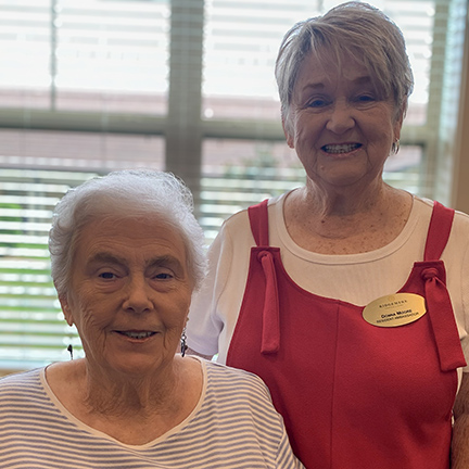 Two senior women, one wearing a gold Resident Ambassador badge, smile brightly at Ridgemere Senior Living.